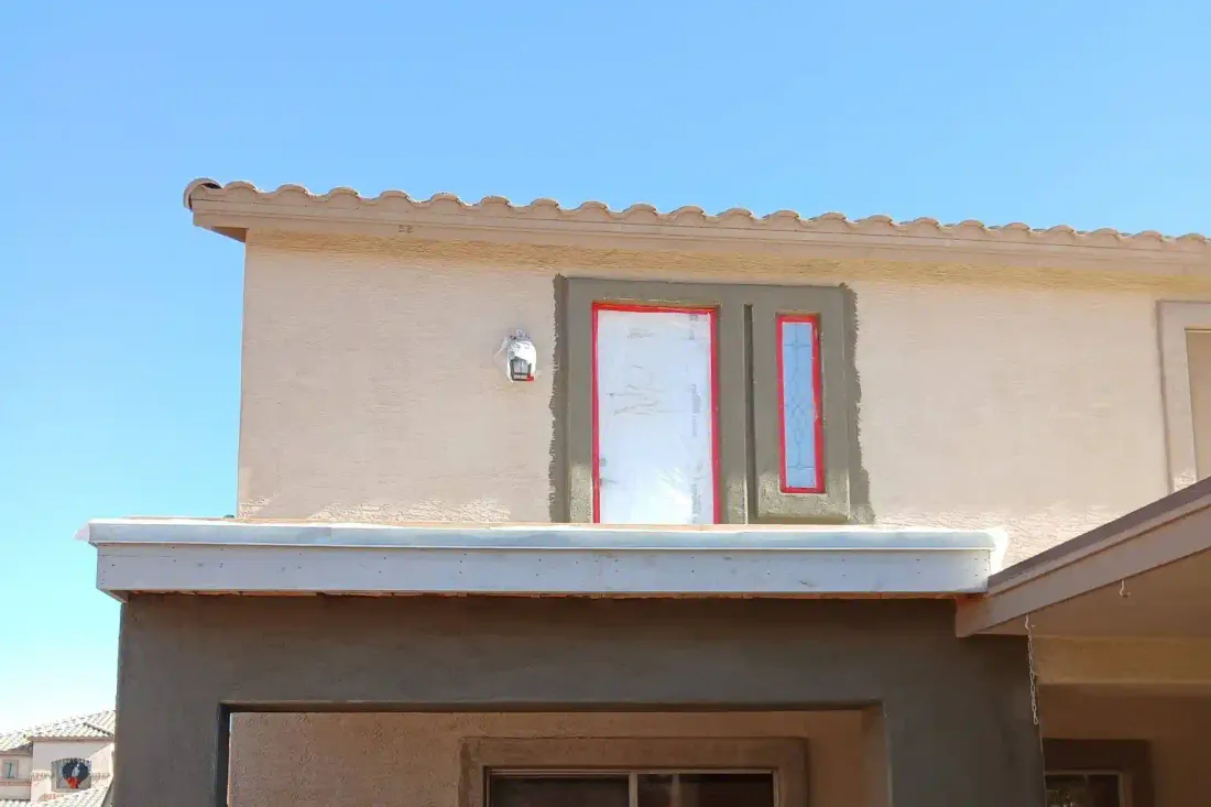 Grey base coat worked around a window and a door opening on a two-storey stucco home, red flashing tape still on both openings and ladders against the wall.