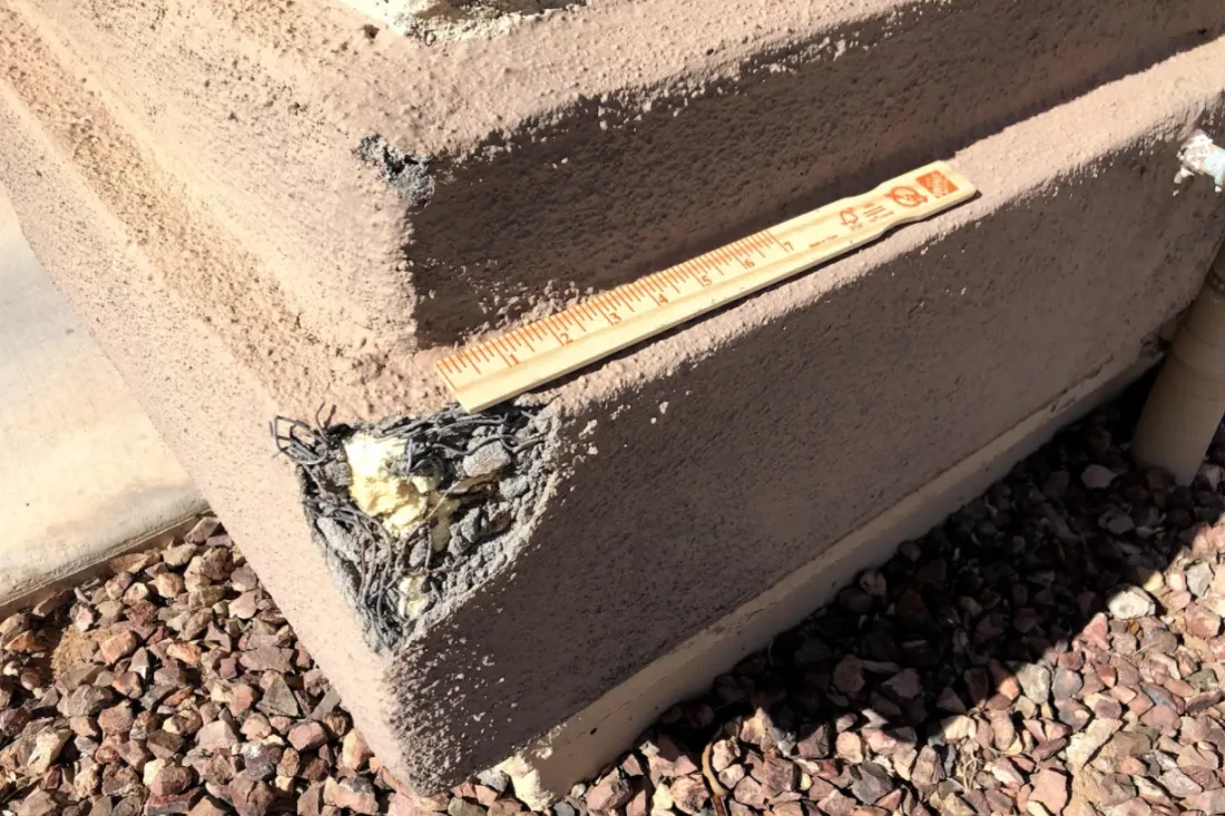 Blown-out corner at the base of a stucco wall with the finish coat broken away, exposing rusted wire lath and crumbling foam, a paint stick laid across the hole for scale.