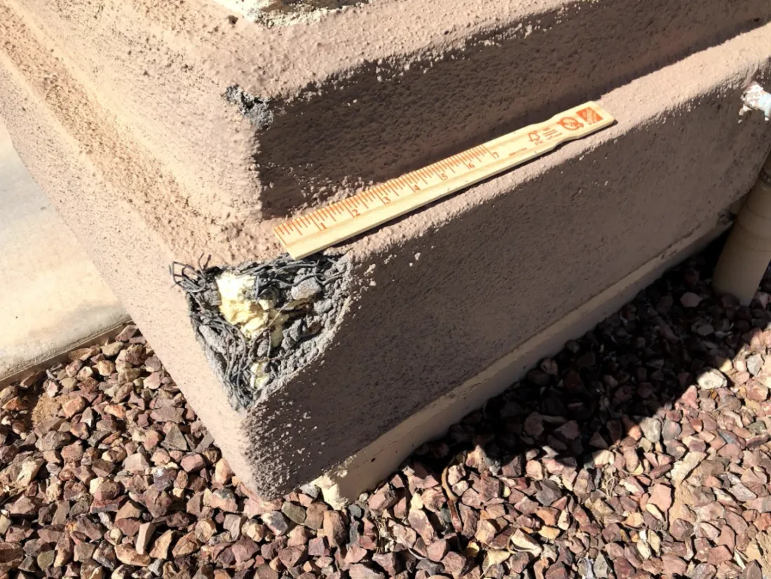 Blown-out corner at the base of a stucco wall with the finish coat broken away, exposing rusted wire lath and crumbling foam, a paint stick laid across the hole for scale.