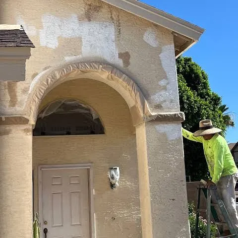 Stucco patches drying in pale rectangles across a tan entry archway, the arch masked with plastic and a plasterer on a ladder working the wall above the door.