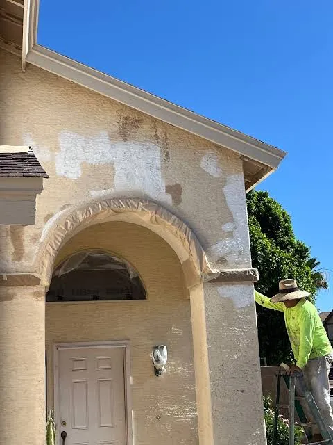 Stucco patches drying in pale rectangles across a tan entry archway, the arch masked with plastic and a plasterer on a ladder working the wall above the door.