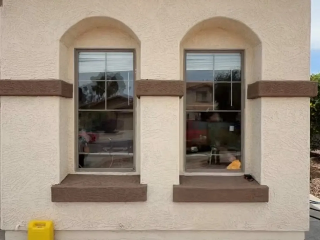 Two arched window openings in a cream stucco wall with brown foam-and-stucco sills and a band course carried across between them.