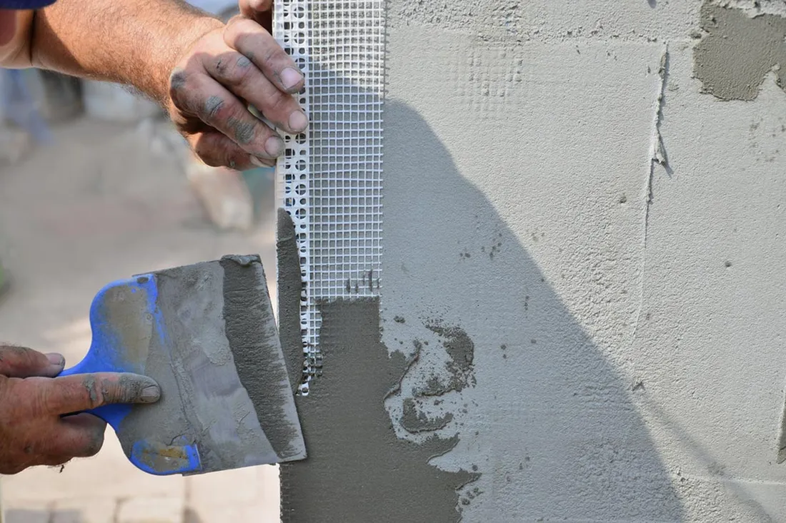 Plasterer bedding fibreglass mesh and a corner bead into wet base coat at a wall corner, working it flat with a blue-handled trowel.