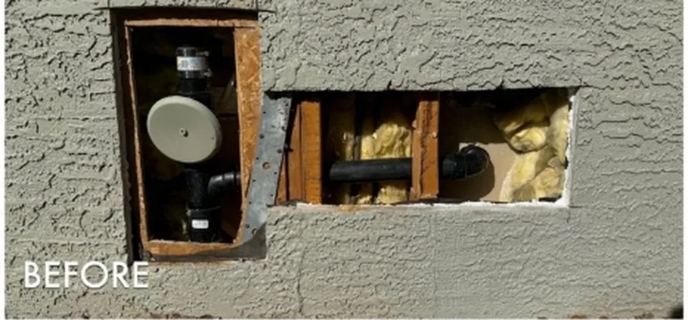 Two rectangular holes cut through a grey stucco wall for plumbing access, exposing black drain pipe, timber studs and yellow fibreglass insulation.