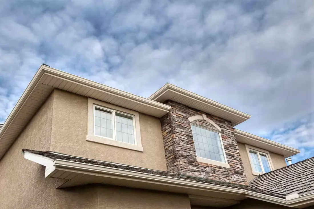 Upper storey of a house seen from below, stucco meeting the soffit and fascia along the roofline beside a stone-veneered gable.