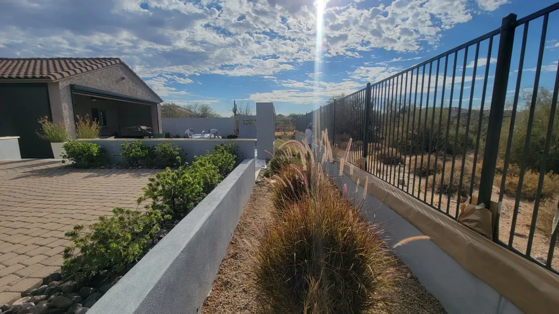 Freshly stuccoed garden and property walls running along a paver patio in a desert back yard, two plasterers working at the far end under hard afternoon sun.