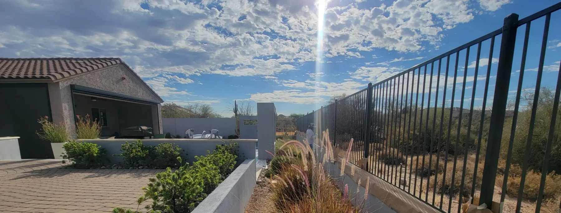 Freshly stuccoed garden and property walls running along a paver patio in a desert back yard, two plasterers working at the far end under hard afternoon sun.
