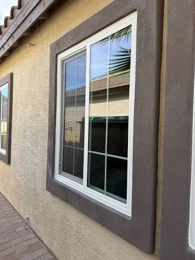 White vinyl window set in a tan stucco wall with a wide dark brown foam-and-stucco trim band run around the opening.